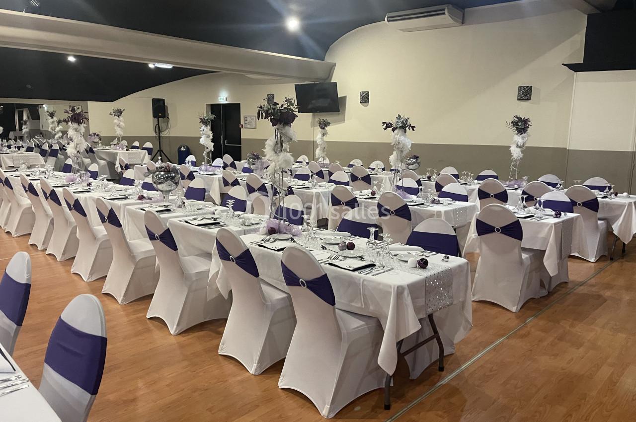 Salle de réception décorée avec des tables alignées, nappes blanches et chaises ornées de housses blanches et nœuds violets.