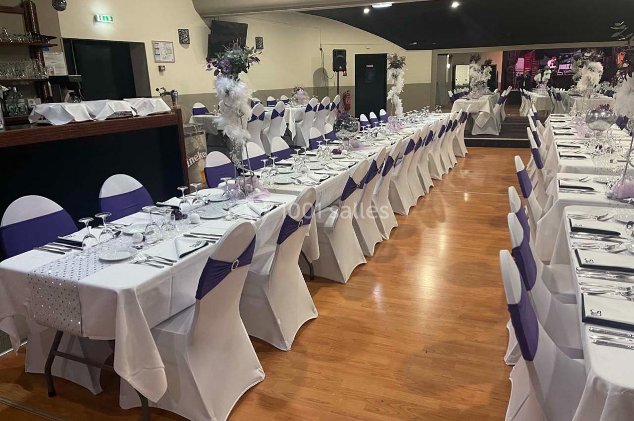 Salle de réception avec tables alignées, nappes blanches, chaises décorées de nœuds violets et décorations florales.