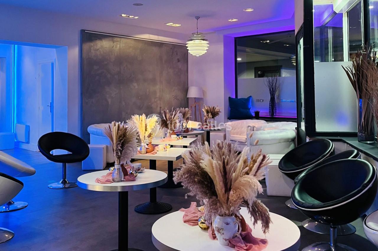 Salle moderne avec fauteuils noirs et blancs, tables décorées de bouquets de fleurs séchées, éclairage tamisé coloré.