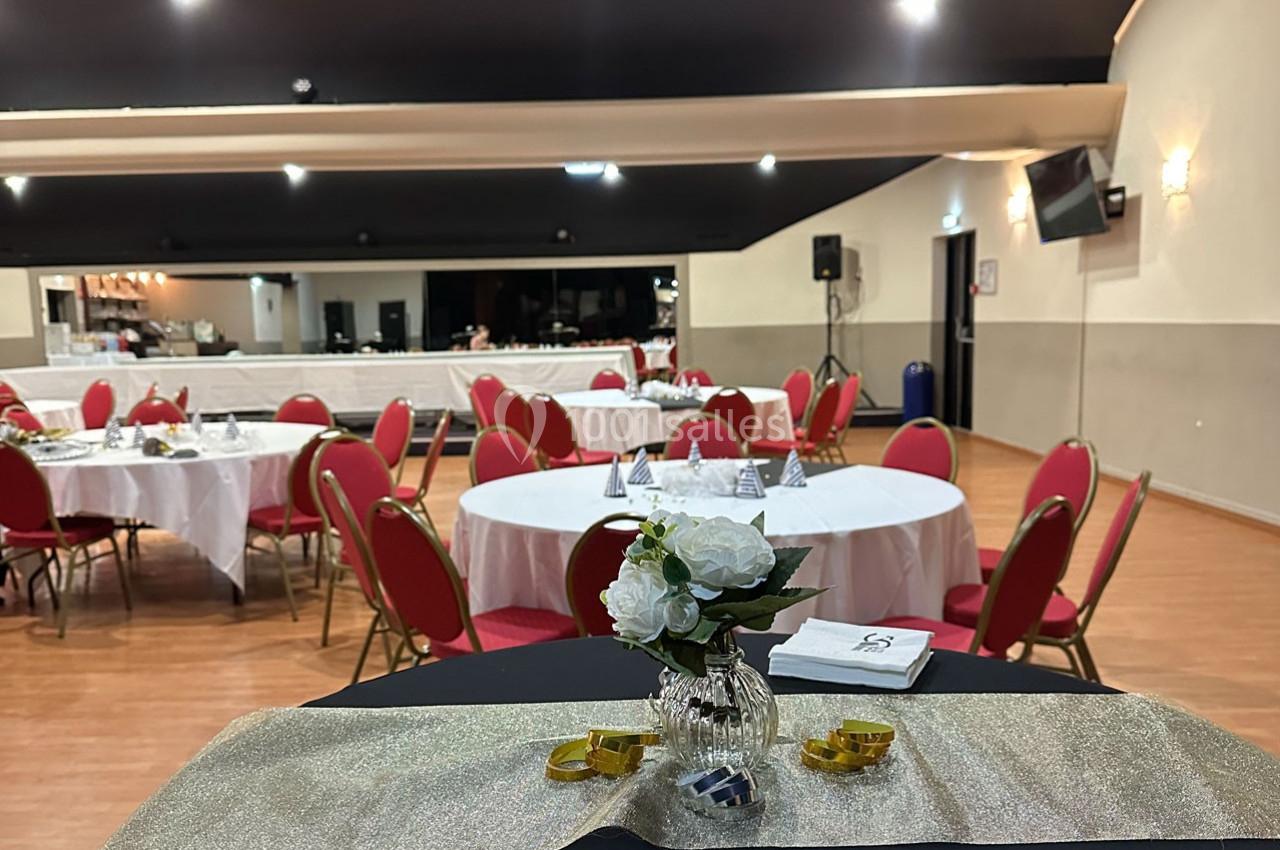 Salle de réception avec tables rondes dressées, chaises rouges et décoration florale sur une table au premier plan.