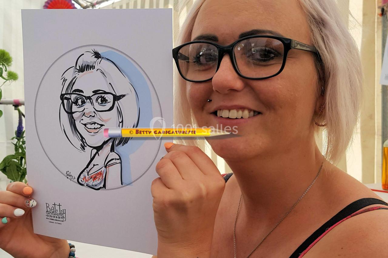 Femme souriante tenant un dessin caricatural d'elle-même, réalisée sur une feuille blanche.