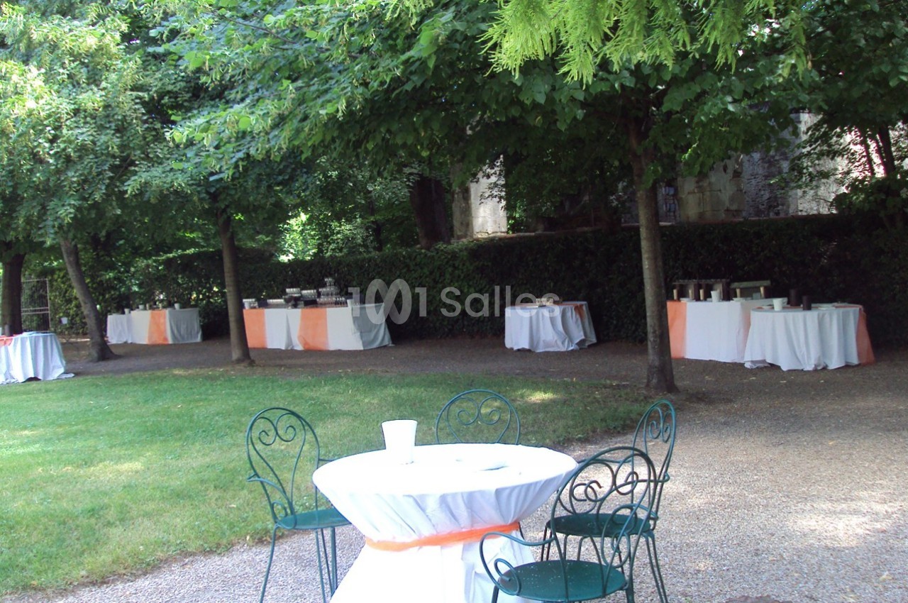 Tables rondes et rectangulaires décorées pour un événement en plein air, disposées sous des arbres dans un jardin.