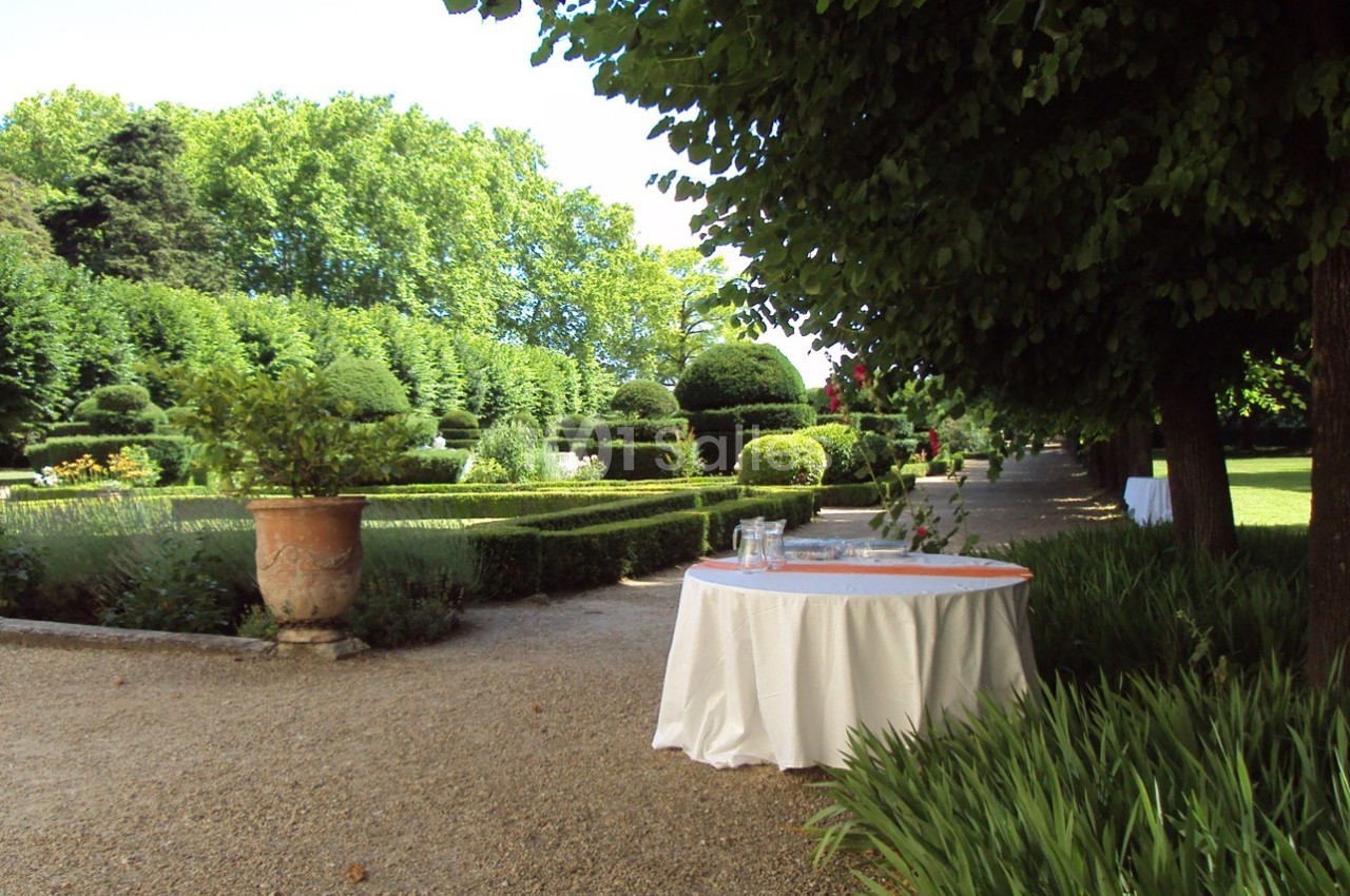 Table ronde avec nappe blanche dans un jardin verdoyant, entourée d'arbres, de haies taillées et d'une allée gravillonnée.