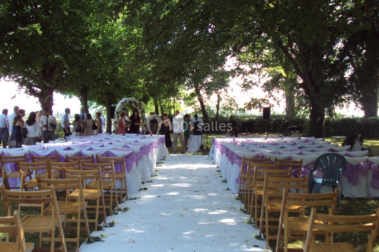 Allée centrale bordée de chaises décorées en violet, menant à une arche pour une cérémonie en plein air sous des arbres.