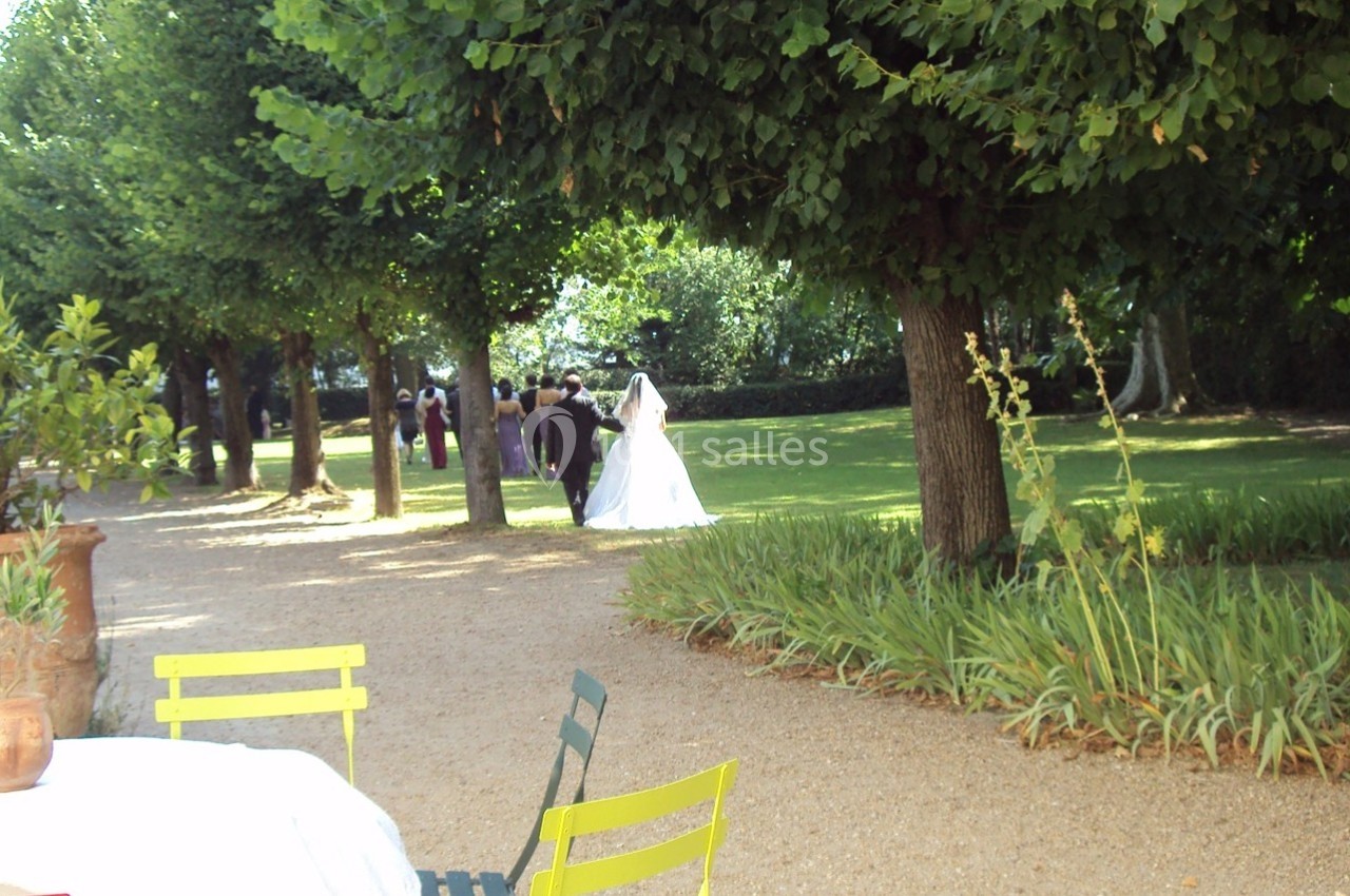 Une mariée en robe blanche marche dans une allée bordée d'arbres, entourée de verdure et de quelques invités.