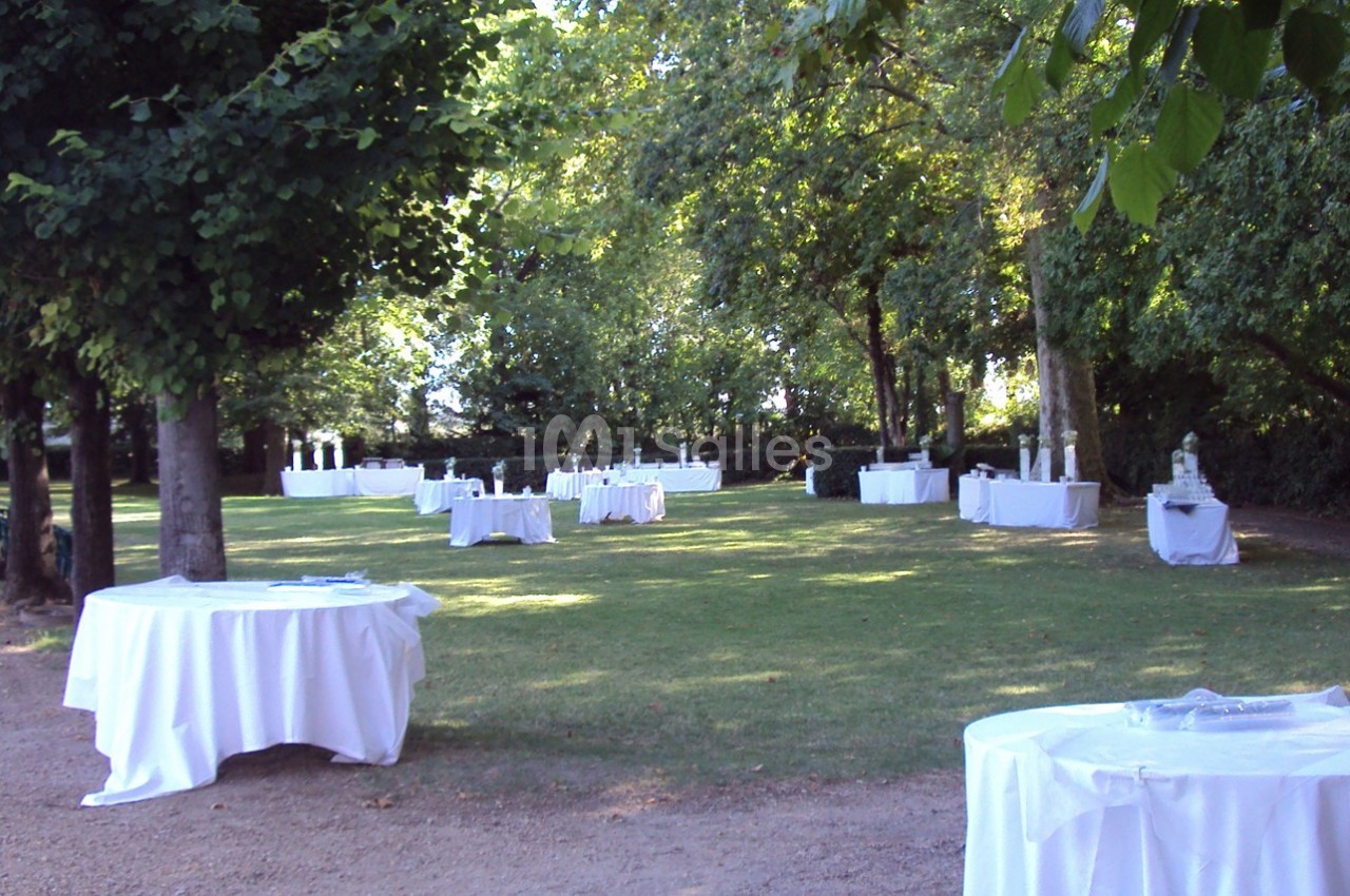 Tables rondes recouvertes de nappes blanches disposées en extérieur dans un jardin ombragé.