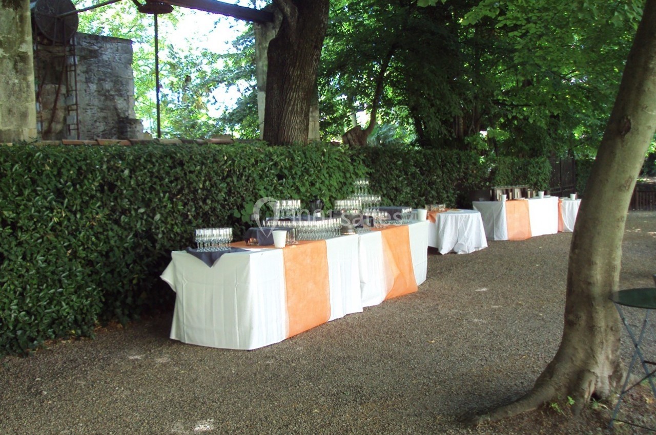 Tables dressées avec nappes blanches et oranges, disposées en extérieur près d'une haie et d'arbres, pour un événement.