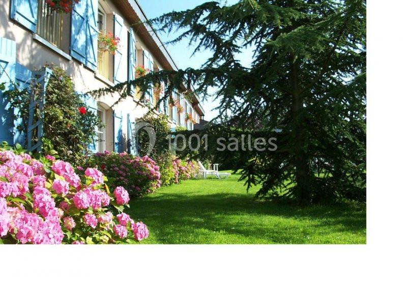 Façade d'une maison avec volets bleus, entourée de massifs d'hortensias roses et d'un grand jardin verdoyant.