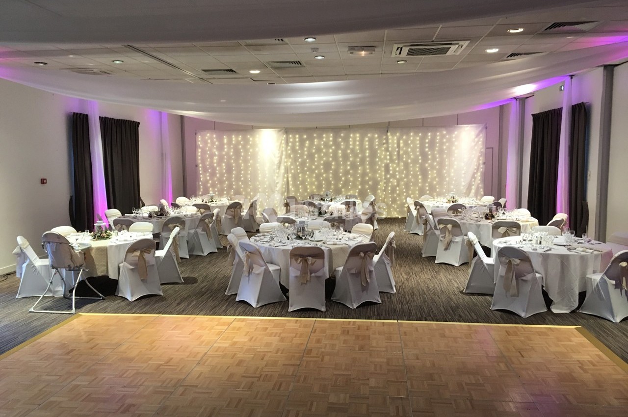 Salle de réception décorée avec des tables rondes dressées, des chaises couvertes et un fond lumineux avec guirlandes.