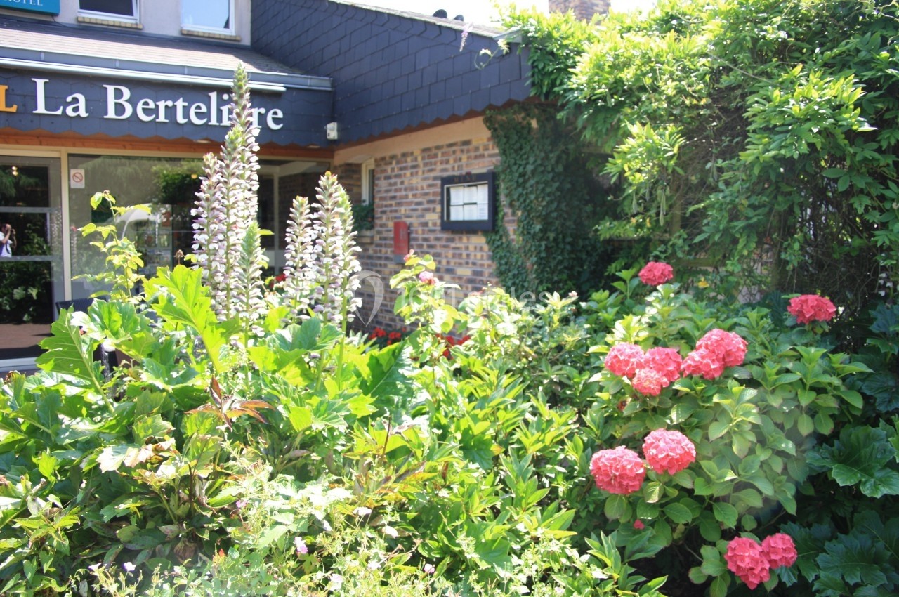 Jardin fleuri avec des hortensias roses et des plantes vertes devant une façade en briques et l'enseigne d'un bâtiment.