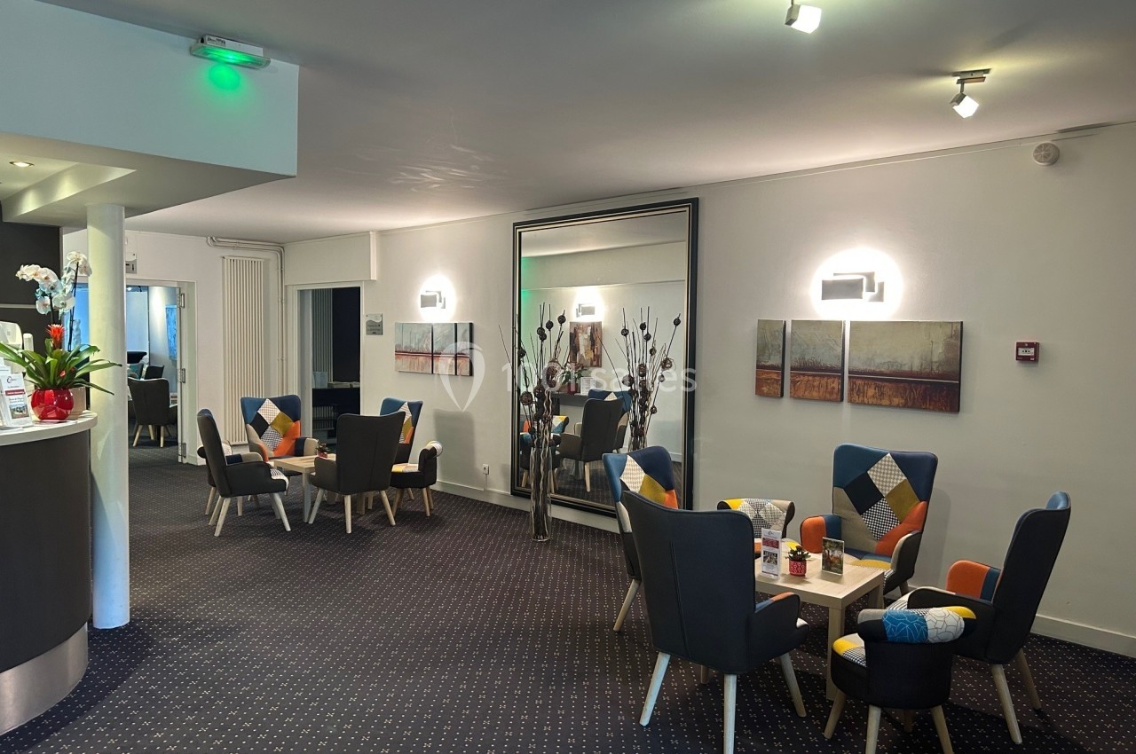 Salon d'hôtel moderne avec fauteuils colorés, tables basses, grand miroir et éclairage chaleureux.