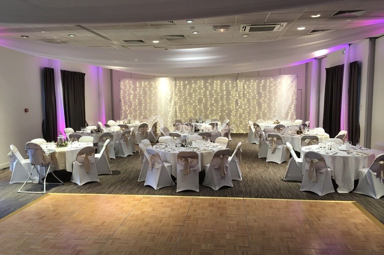 Salle de réception décorée avec des tables rondes dressées, des chaises couvertes et un fond lumineux avec guirlandes.