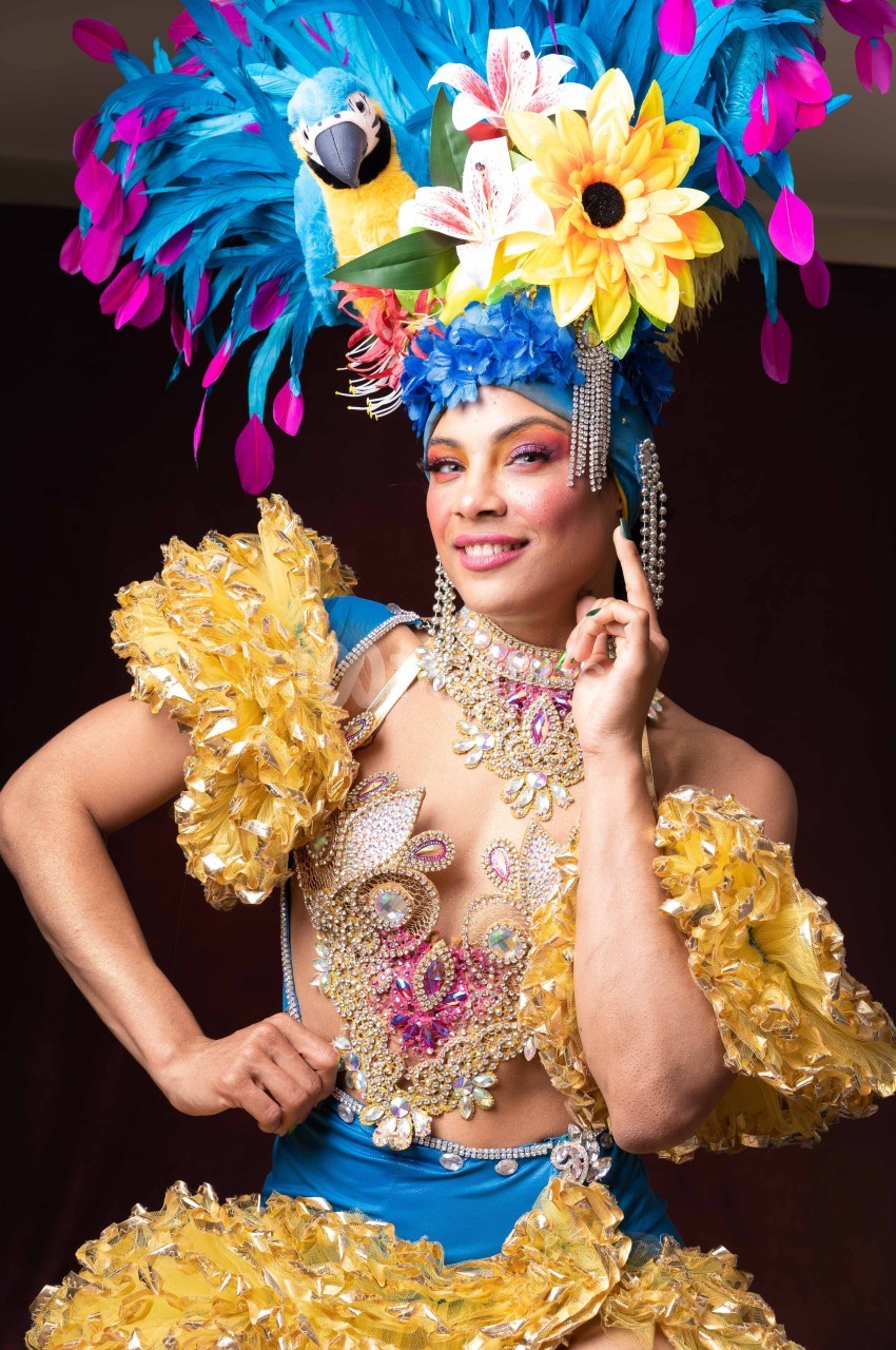 Personne en costume coloré avec plumes, perles et fleurs, posant devant un fond sombre.