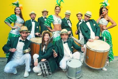 Trois hommes souriants en tenue de capoeira avec des drapeaux brésiliens, posant dans un parc ensoleillé.