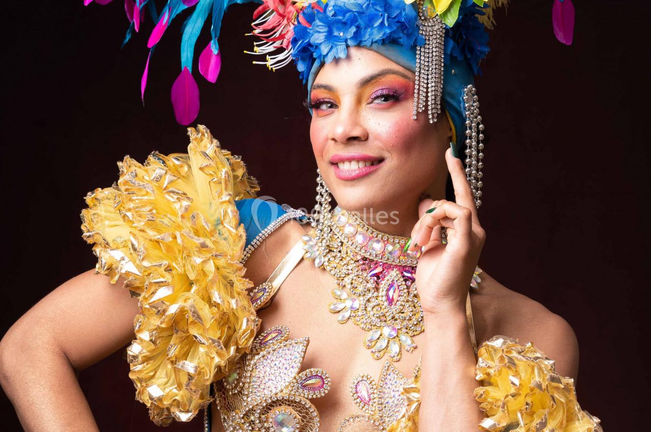 Personne souriante en costume coloré avec plumes, perles et ornements, posant sur un fond sombre.