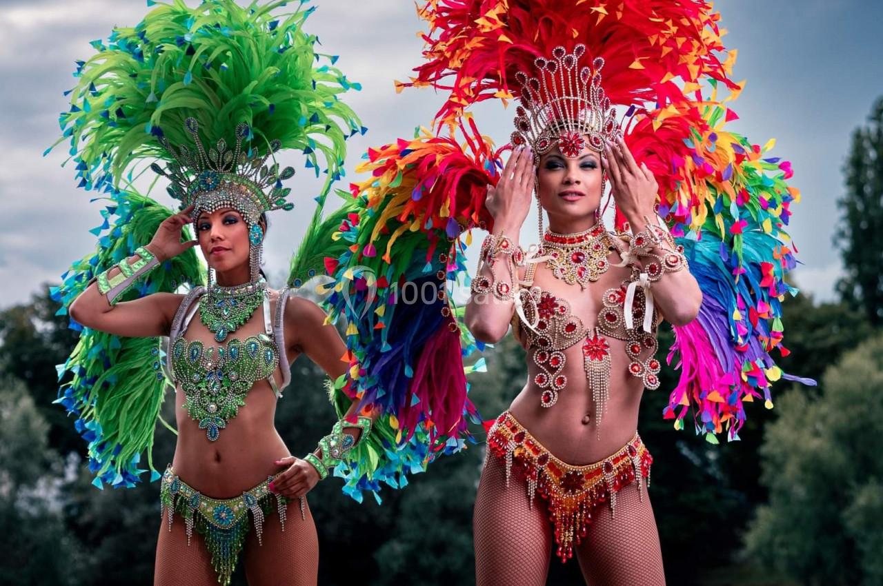 Deux danseuses en costumes colorés et ornés de plumes posent en extérieur devant un paysage arboré.