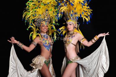 Trois hommes souriants en tenue de capoeira avec des drapeaux brésiliens, posant dans un parc ensoleillé.