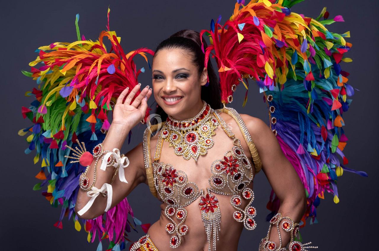 Danseuse en costume coloré avec plumes multicolores et ornements scintillants, posant sur un fond sombre.