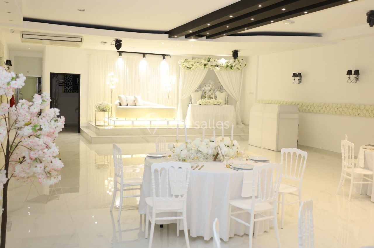 Salle de réception lumineuse décorée de blanc avec table dressée, fleurs et estrade ornée pour un événement.
