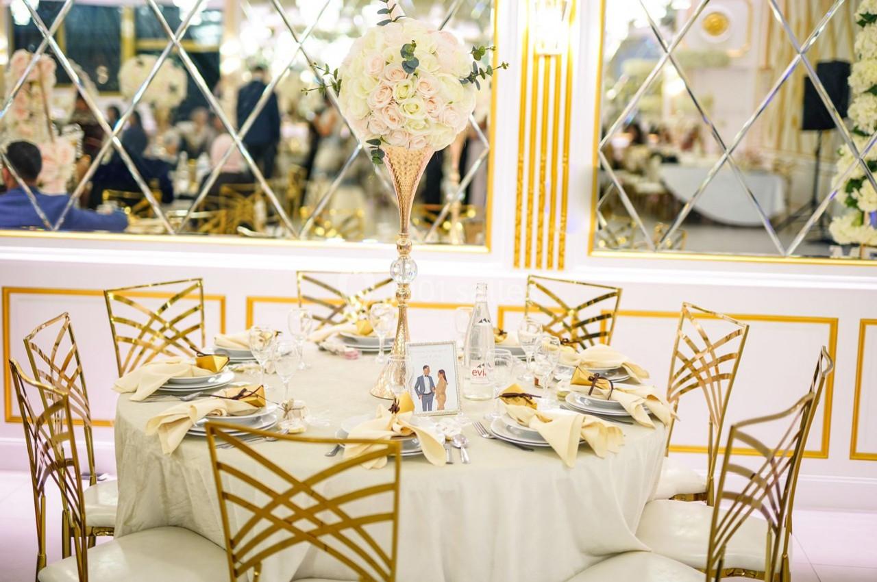 Table élégamment dressée avec nappes claires, chaises dorées et centre de table floral dans une salle ornée de miroirs.