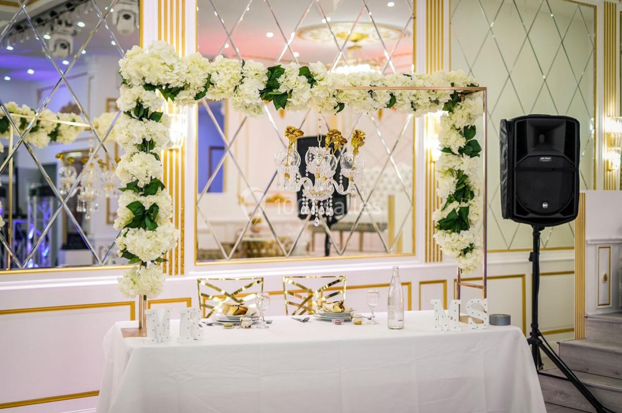 Table décorée pour un mariage avec arche florale blanche, chandelier suspendu et lettres ’Mr & Mrs’.