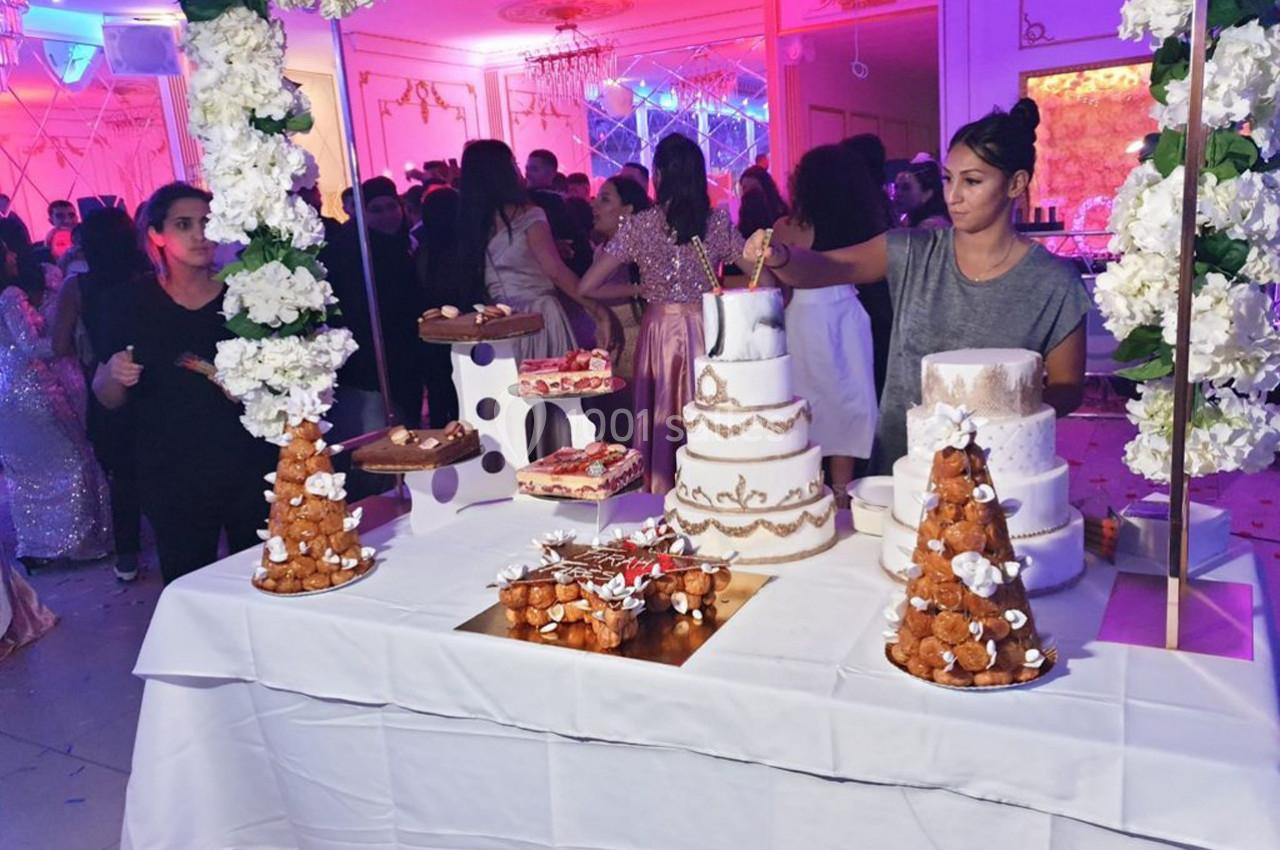 Table de desserts décorée avec des gâteaux, pièces montées et fleurs blanches, dans une salle de réception festive.