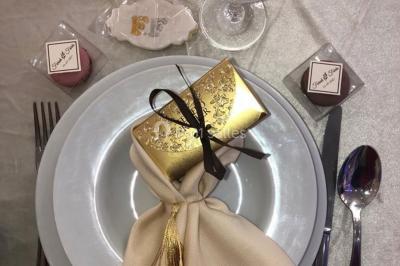 Table dressée avec une assiette blanche, un cadeau doré, une serviette beige nouée et des macarons en décoration.