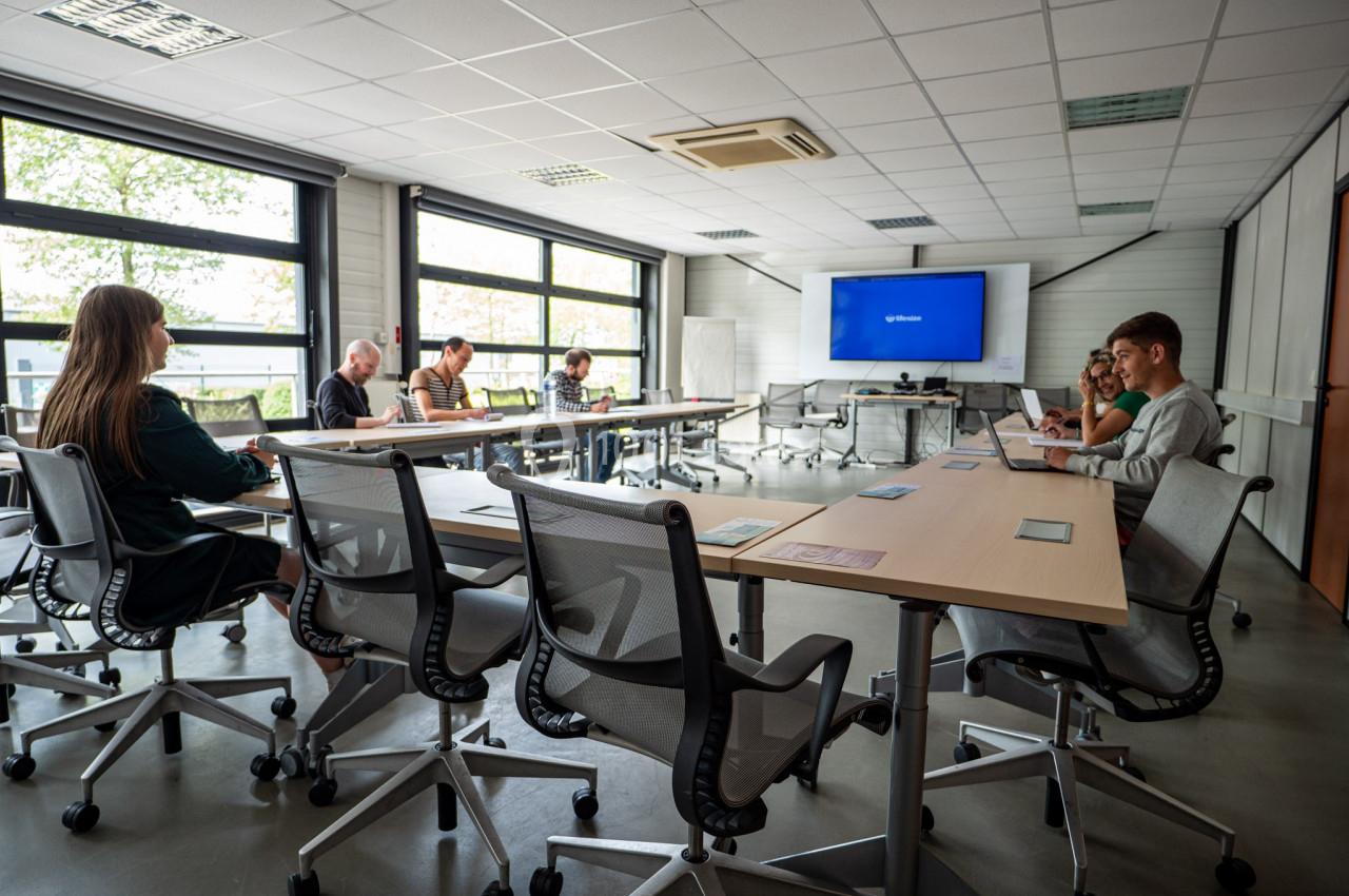 Salle de réunion lumineuse avec plusieurs personnes assises autour de tables, certaines utilisant des ordinateurs.