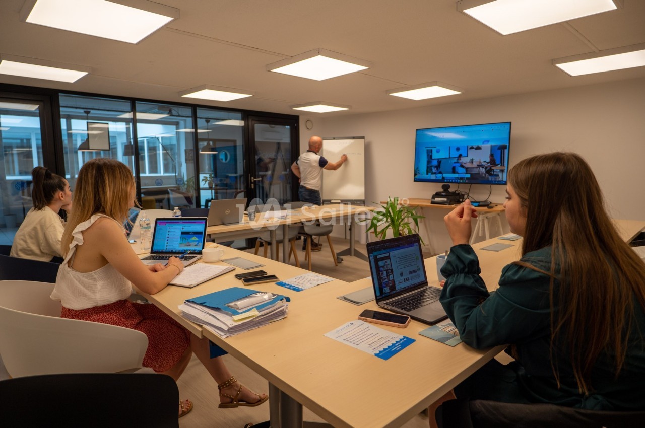 Un formateur écrit sur un tableau blanc devant trois personnes travaillant sur des ordinateurs dans une salle de réunion.