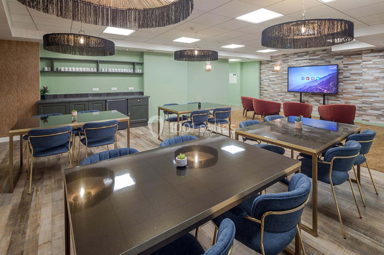 Salle moderne avec tables noires, chaises bleues, étagères murales, et écran mural dans un espace lumineux et décoré.