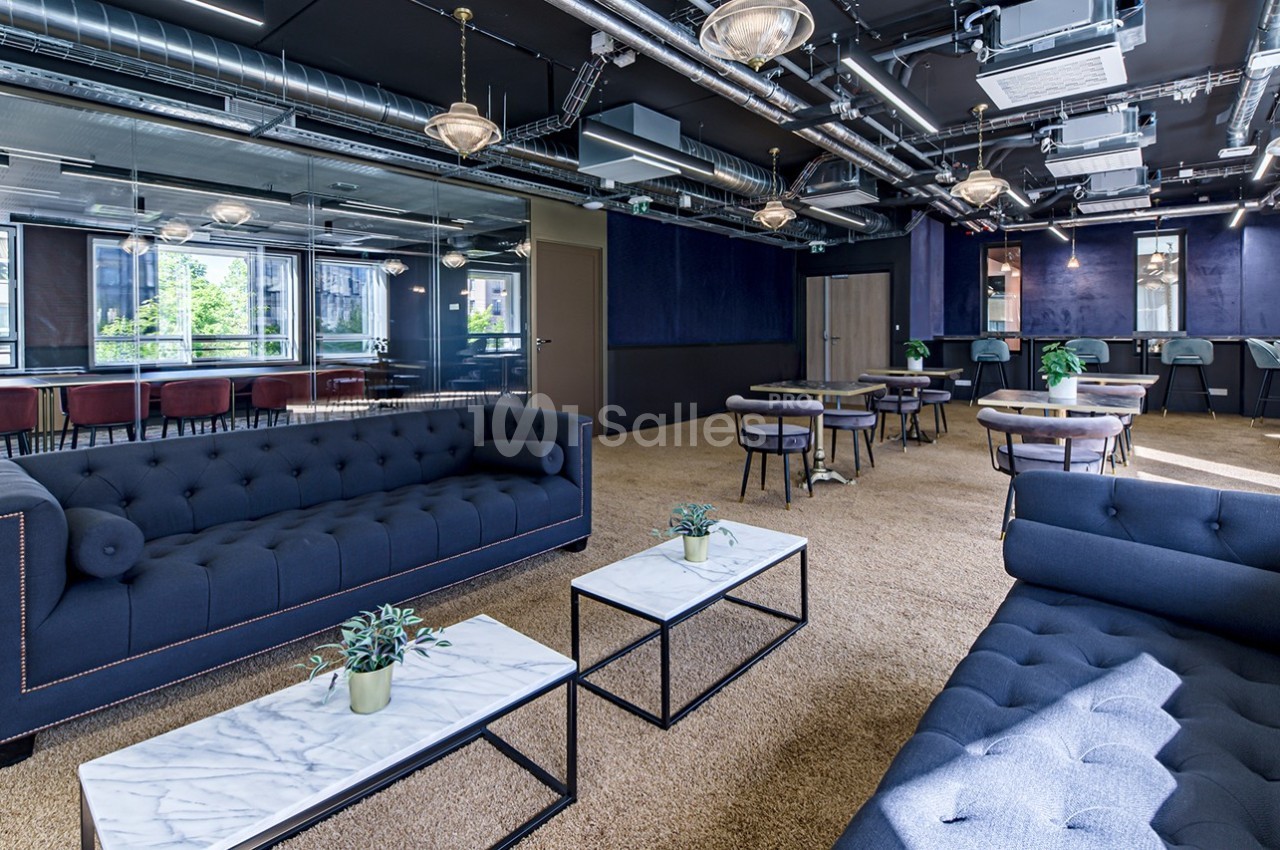 Espace intérieur moderne avec canapés bleus, tables basses en marbre, et grandes baies vitrées laissant entrer la lumière.