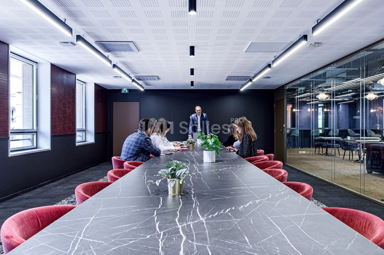 Salle de réunion moderne avec une grande table en marbre, des chaises rouges et un groupe de personnes en discussion.