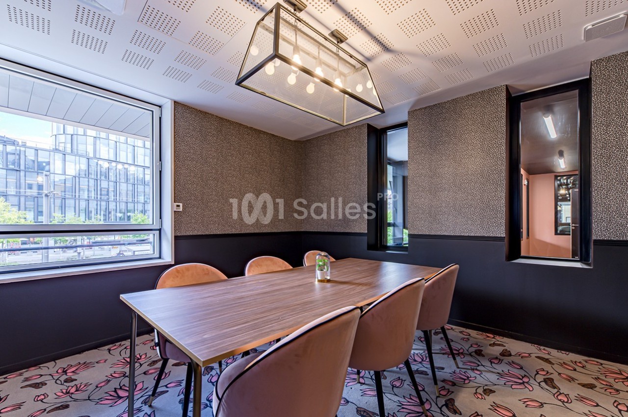 Salle de réunion lumineuse avec table en bois, chaises roses, suspension moderne et vue sur des immeubles extérieurs.