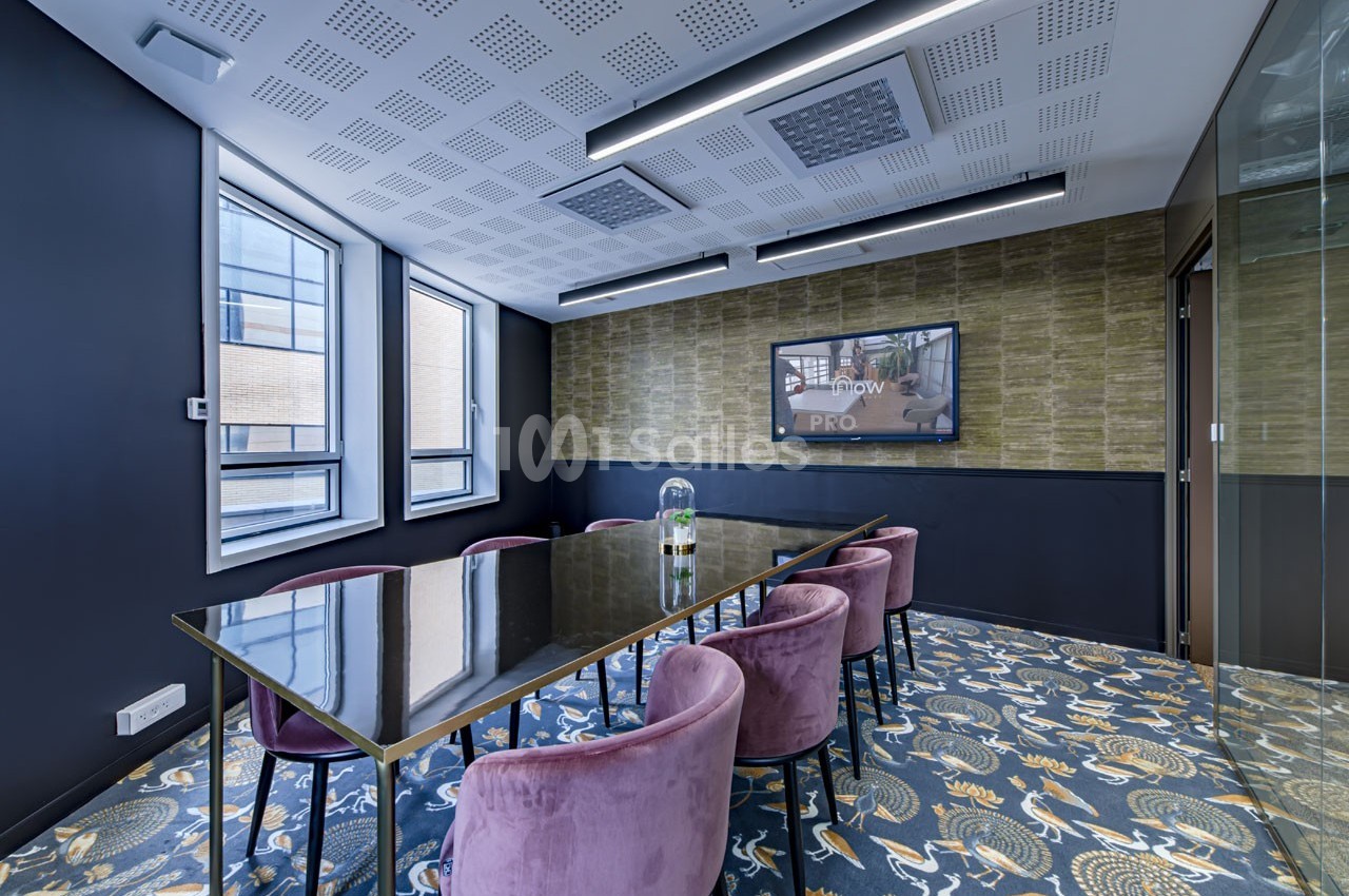 Salle de réunion moderne avec table en verre, chaises violettes, écran mural et fenêtres laissant entrer la lumière…