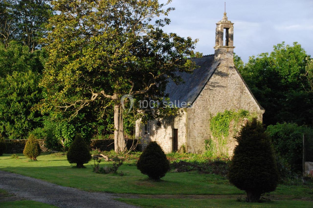 Petite chapelle en pierre entourée de verdure, avec un arbre imposant et des buissons dans un jardin.