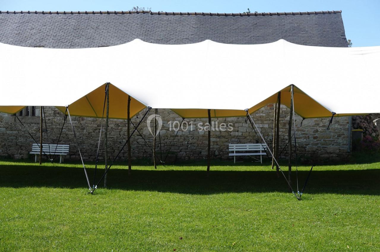 Tente blanche sur une pelouse verte, soutenue par des structures métalliques, avec un mur en pierre en arrière-plan.
