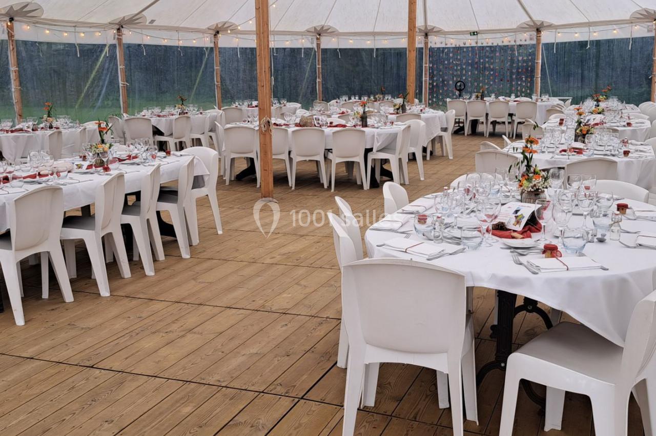 Tables rondes et rectangulaires dressées pour un événement dans une tente avec sol en bois et chaises blanches.
