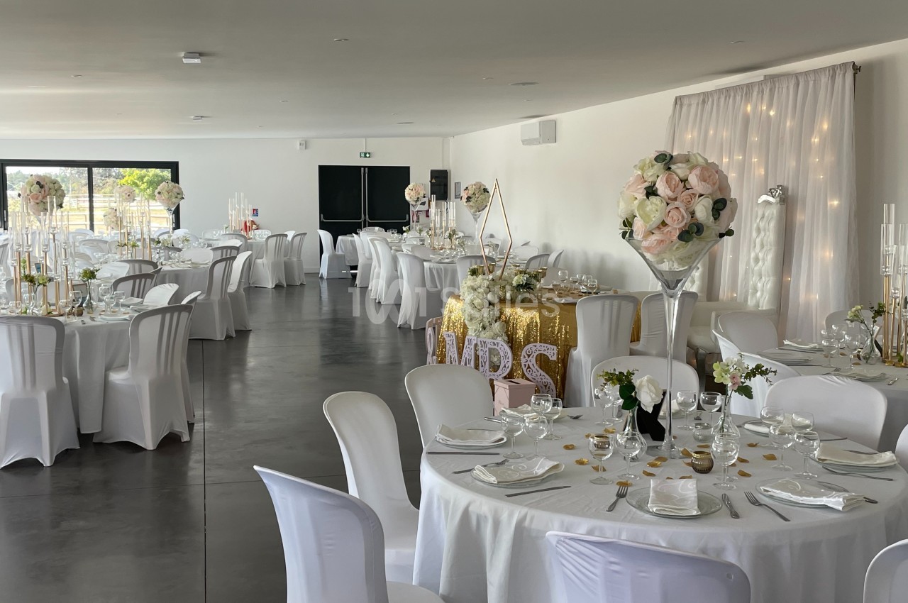 Salle de réception décorée pour un mariage, avec tables dressées, nappes blanches, fleurs et éclairage tamisé.
