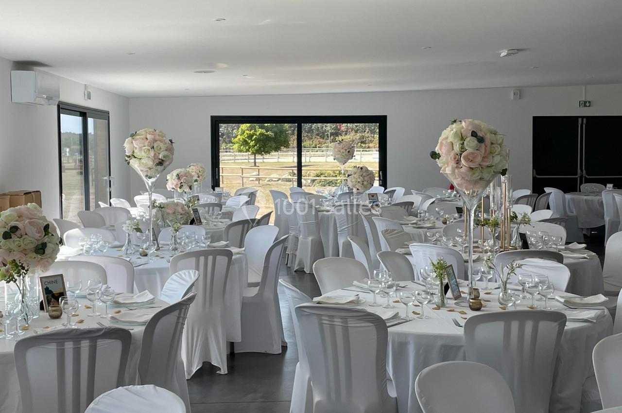 Salle de réception décorée avec des tables rondes, nappes blanches, chaises houssées et centres de table fleuris.