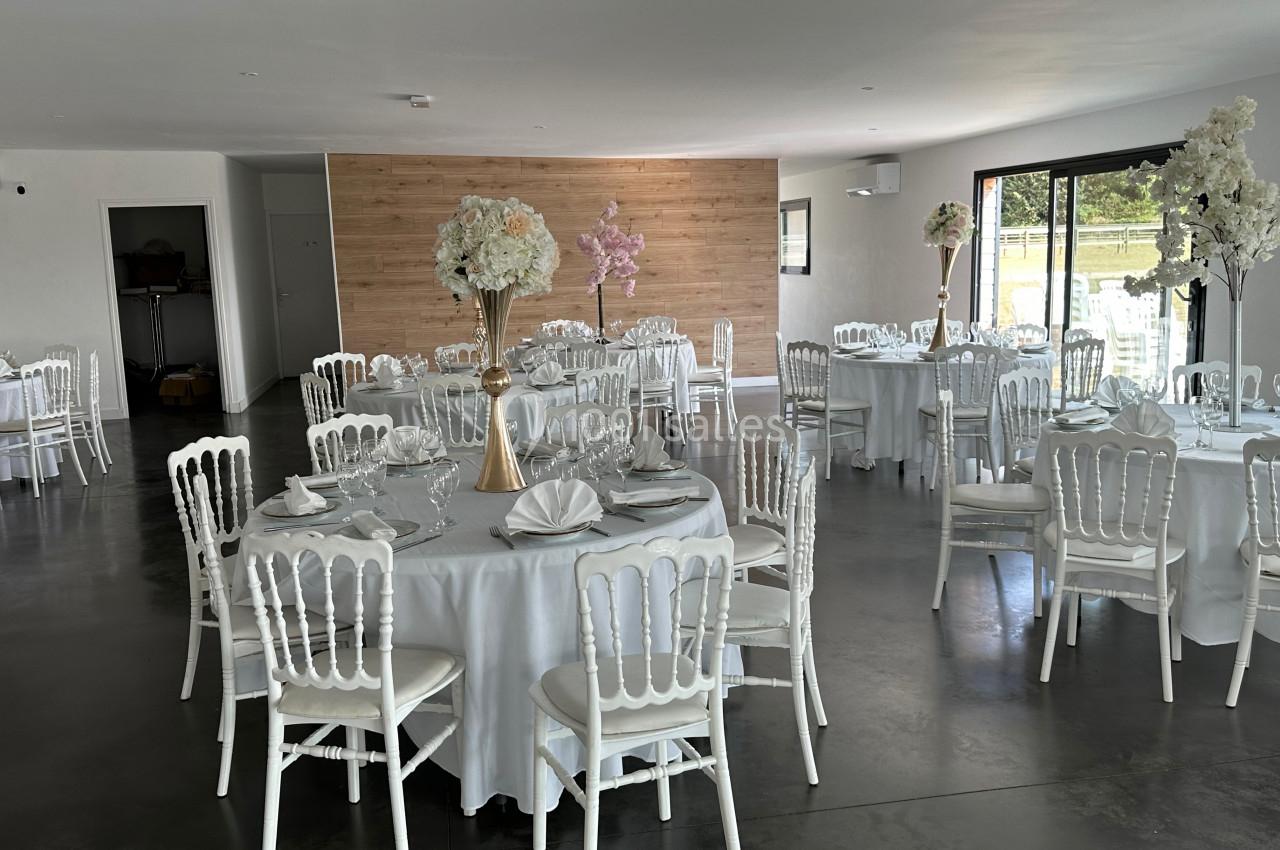 Salle de réception lumineuse avec tables rondes dressées, nappes blanches et chaises blanches, décor floral élégant.