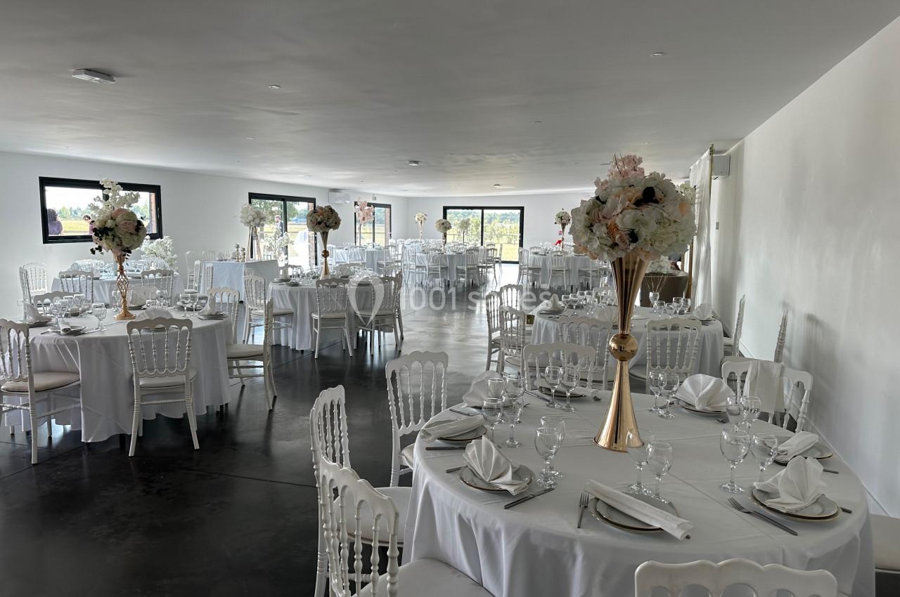 Salle de réception lumineuse avec tables rondes dressées, nappes blanches et centres de table fleuris.