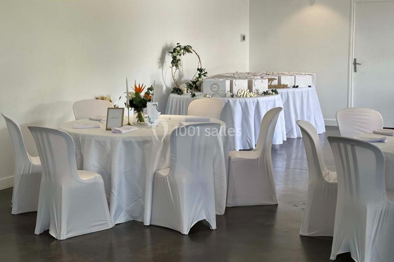 Salle décorée pour un événement avec tables rondes nappées de blanc, chaises assorties et buffet en arrière-plan.