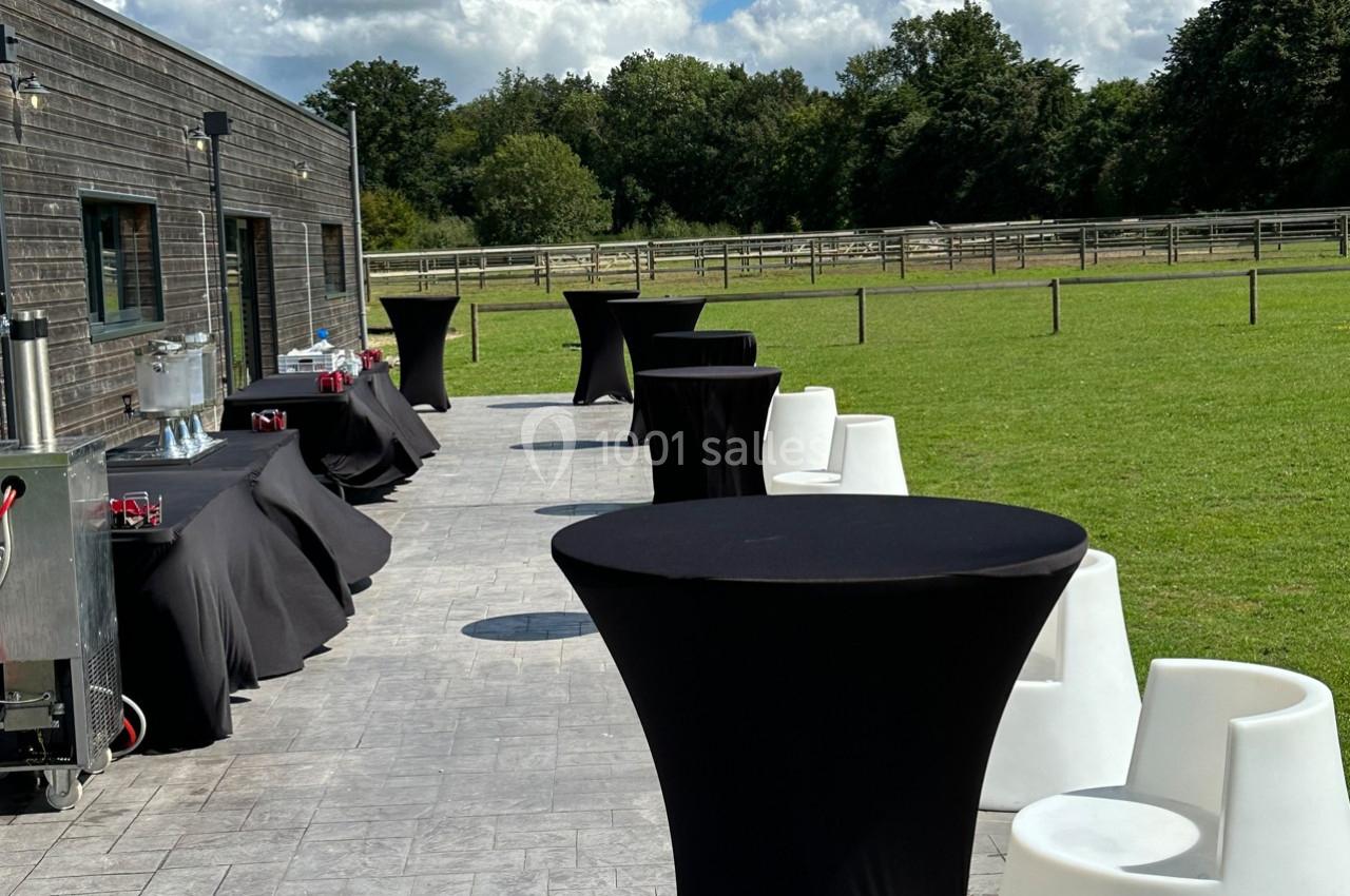 Tables hautes noires et chaises blanches disposées sur une terrasse ensoleillée, avec vue sur un grand espace vert.