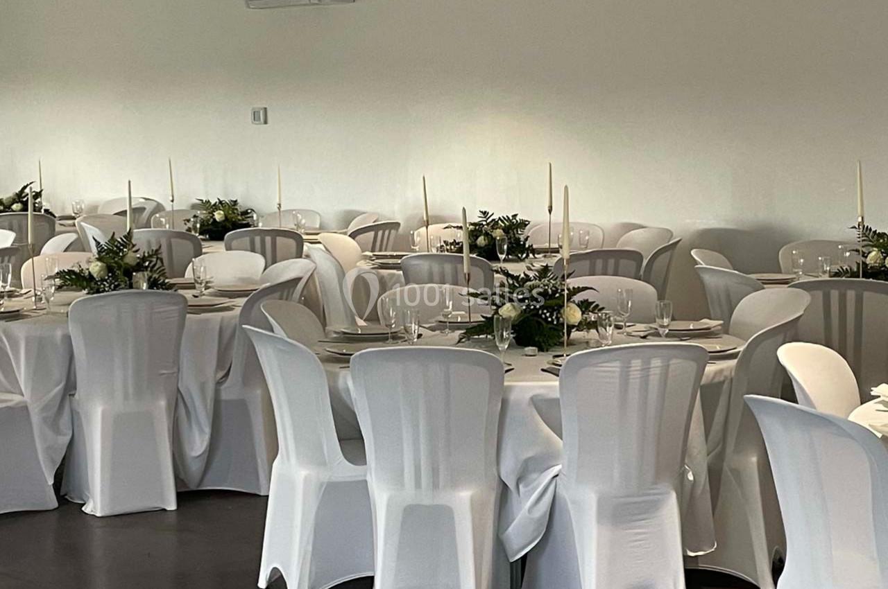 Salle de réception avec des tables rondes dressées, nappes blanches, chaises assorties et décorations florales.
