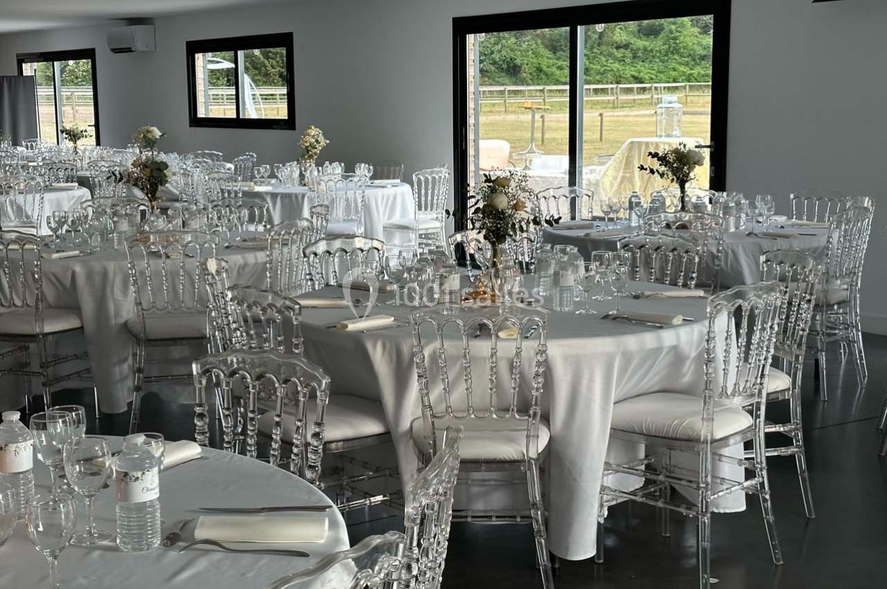 Salle de réception lumineuse avec des tables rondes dressées de nappes blanches et des chaises transparentes.