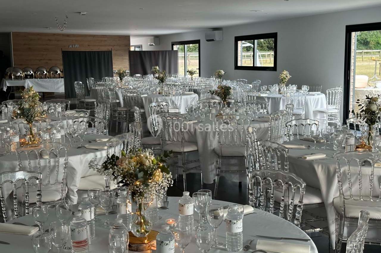 Salle de réception décorée avec des tables rondes, nappes blanches, chaises transparentes et centres de table fleuris.
