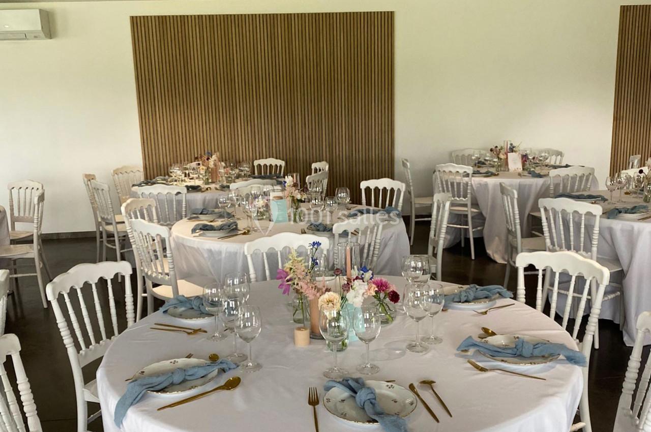 Salle de réception avec tables rondes dressées, nappes blanches, couverts dorés et décorations florales pastel.