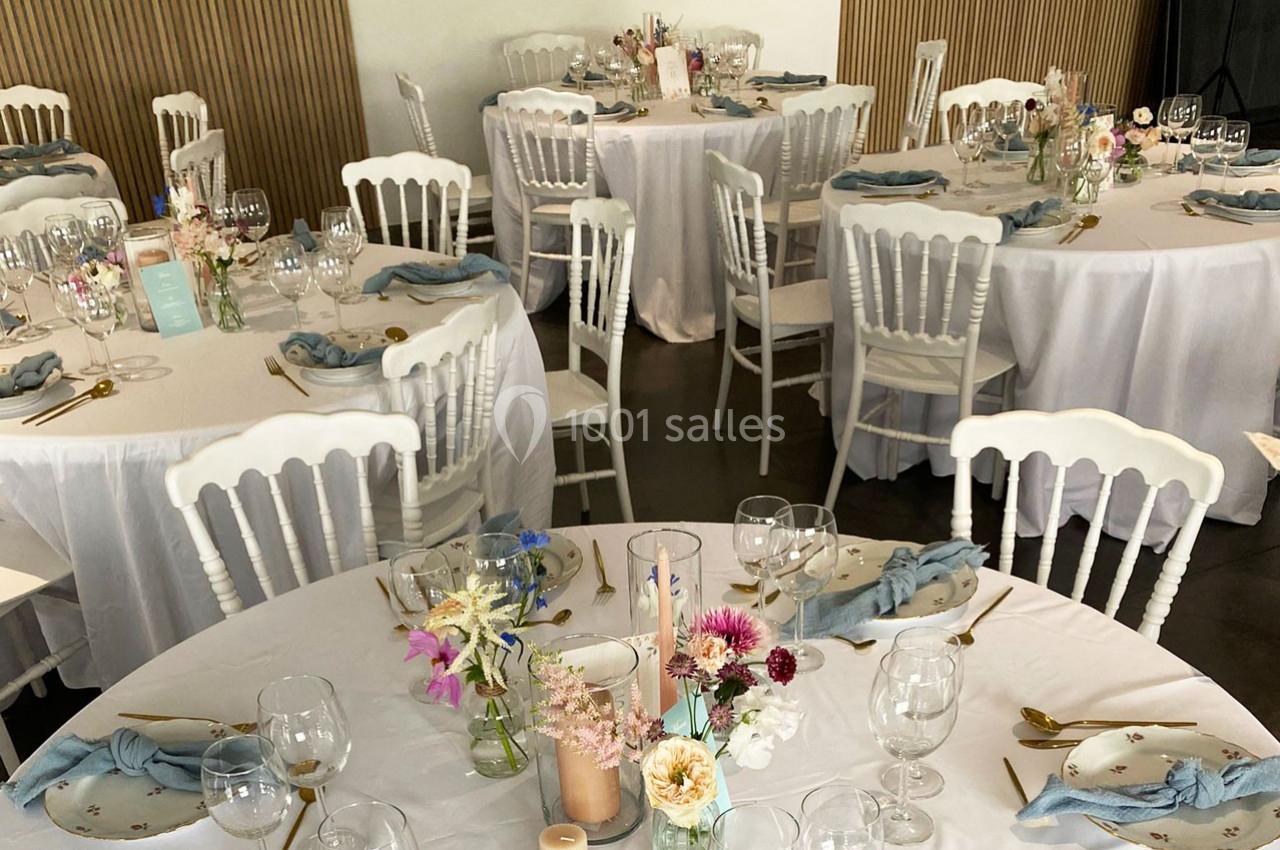 Tables rondes décorées pour un événement, avec nappes blanches, fleurs pastel, serviettes bleues et couverts dorés.