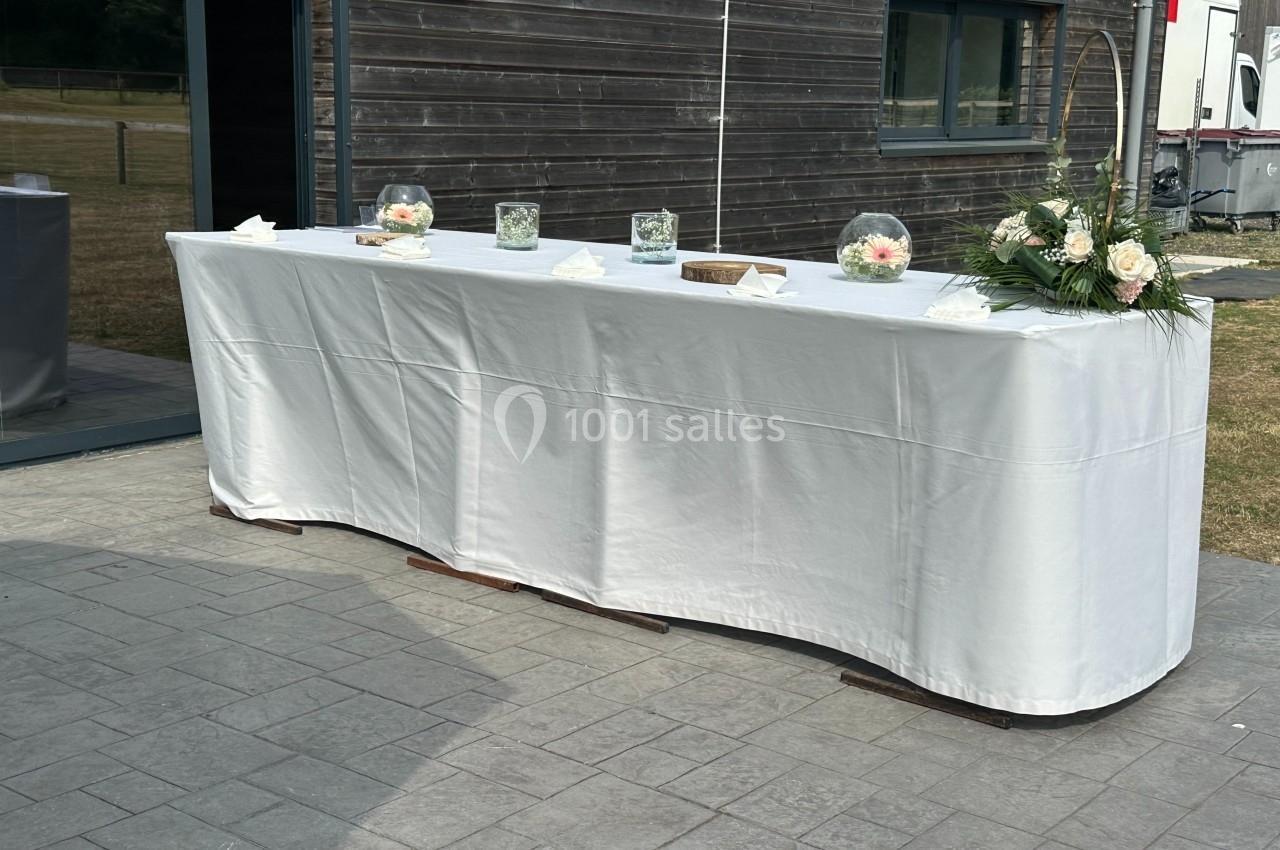 Table recouverte d'une nappe blanche, décorée de fleurs et de vases, placée devant un bâtiment en bois.