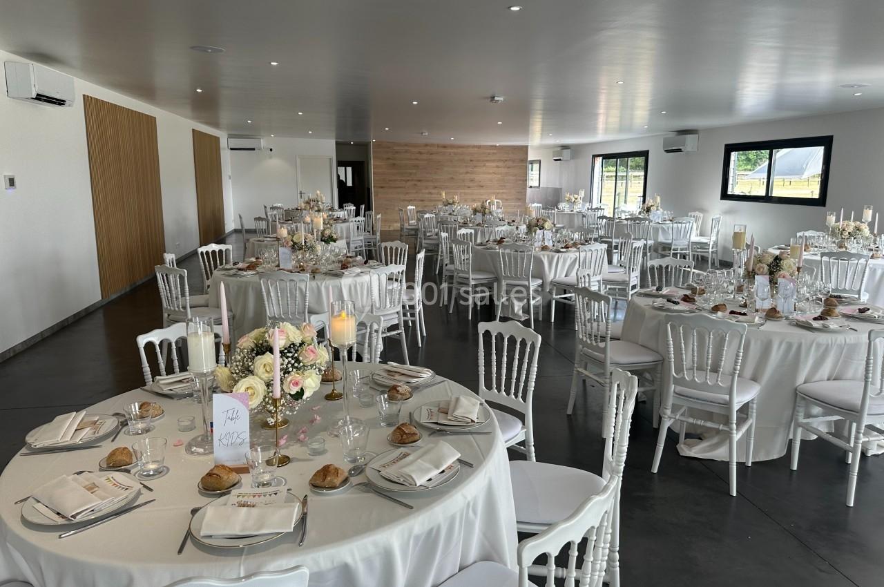 Salle de réception décorée pour un mariage, avec tables rondes dressées, nappes blanches et arrangements floraux.